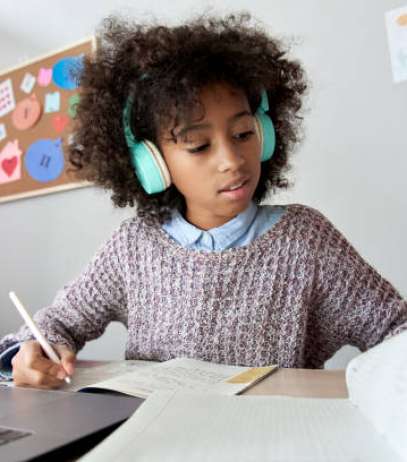 Smart african mixed race kid girl wearing headphones listening lesson learning online computer doing homework at home. Cute black child studying distance school education writing notes sitting at desk